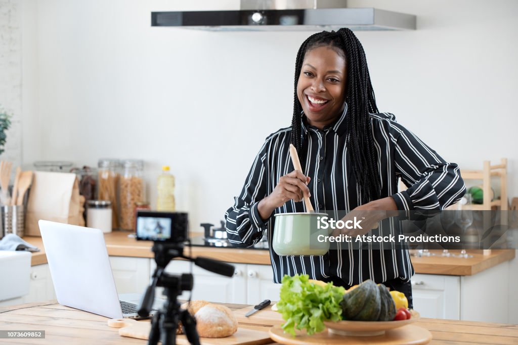 African american woman cooking vlogger recording video in kitchen at home Happy african american woman vlogger broadcasting live video online while cooking food in kitchen at home Cooking Stock Photo