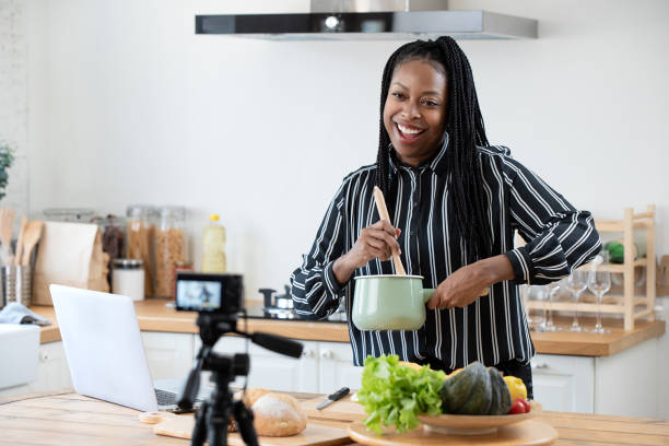 African american woman cooking vlogger recording video in kitchen at home Happy african american woman vlogger broadcasting live video online while cooking food in kitchen at home cooking show stock pictures, royalty-free photos & images