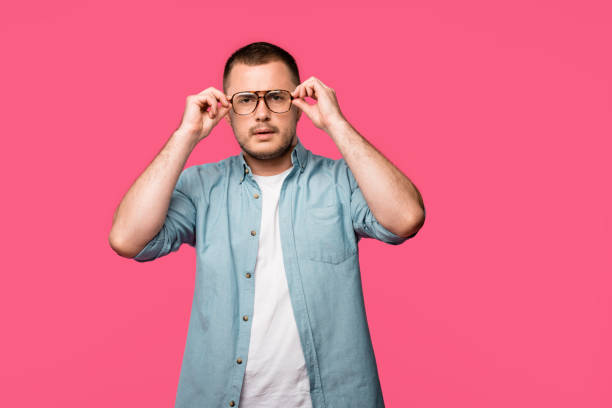 istock serious young man adjusting eyeglasses and looking at camera isolated on pink 1073634740