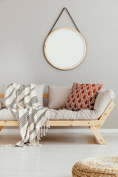 round mirror in wooden frame on the wall of bright beige living room with comfortable sofa with patterned pillow and striped blanket, real photo - wall mirror imagens e fotografias de stock