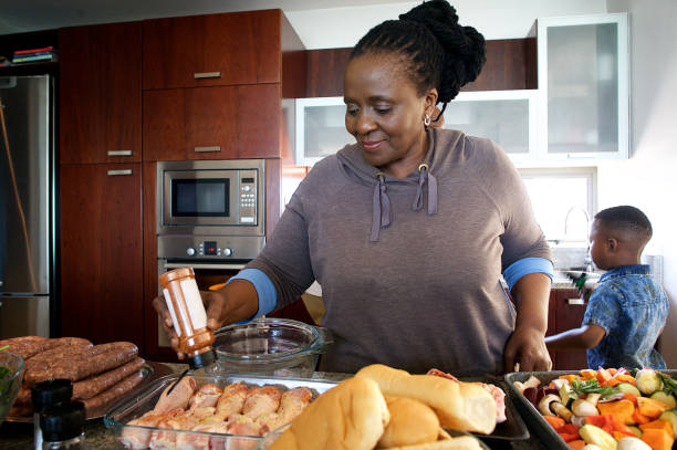 Senior African Woman in kitchen seasoning chicken with grandson in the background Senior African Woman in the kitchen pouring salt seasoning on chicken with grandson in the background Strand Cape Town South Africa salt seasoning stock pictures, royalty-free photos & images
