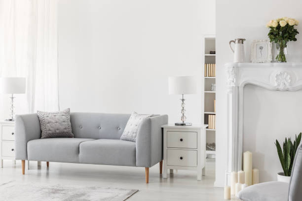istock Luxury grey sofa with pillows between two white wooden commodes with stylish lamps, real photo with copy space on wall 1073680326