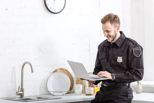 istock handsome man in police uniform smiling and using laptop at kitchen 1073686204