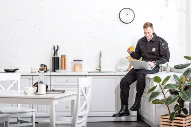istock Young policeman sitting on table, drinking orange juice and using laptop at kitchen 1073686212