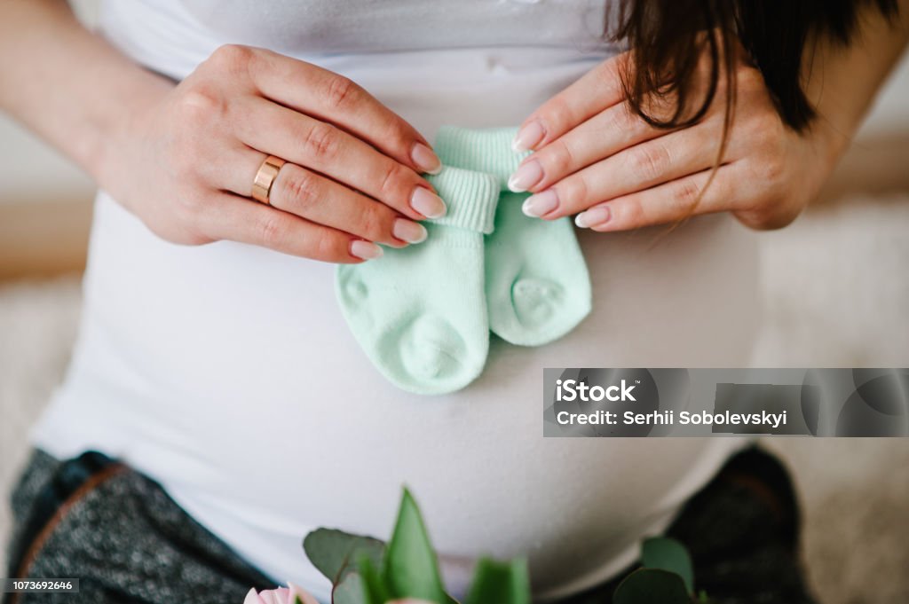 Cropped image pregnant woman holding socks for a baby on her belly stomach. Pregnant hugging tummy at home. Motherhood concept. Baby Shower. Baby - Human Age Stock Photo