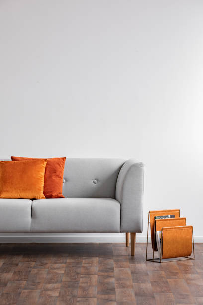 istock Vertical view of grey comfortable couch with orange pillows and newspaper rack next to it, real photo with copy space on the white wall 1073694770