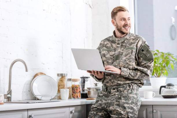istock smiling army soldier using laptop at kitchen and looking away 1073700134