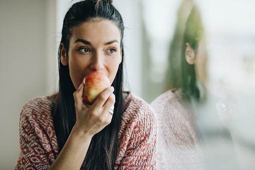 Young beautiful woman eating fresh apple and looking through window.