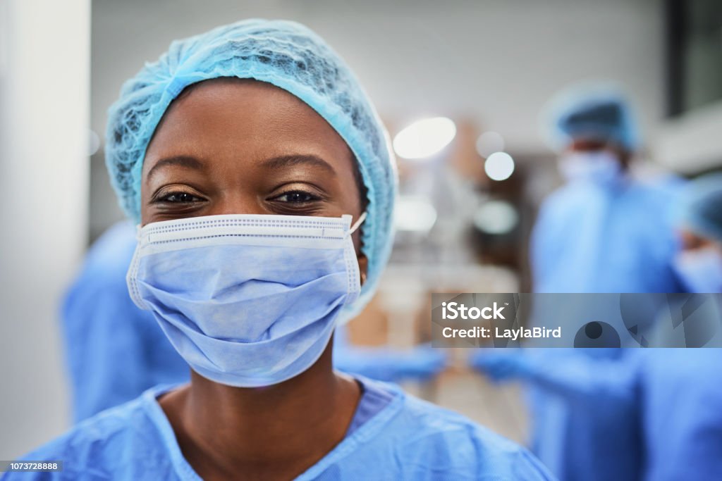 We're willing to go the extra mile Cropped portrait of a young female nurse standing in the OR with her colleagues in the background Nurse Stock Photo