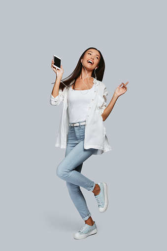 Full length of young Asian woman dancing and smiling while standing against grey background