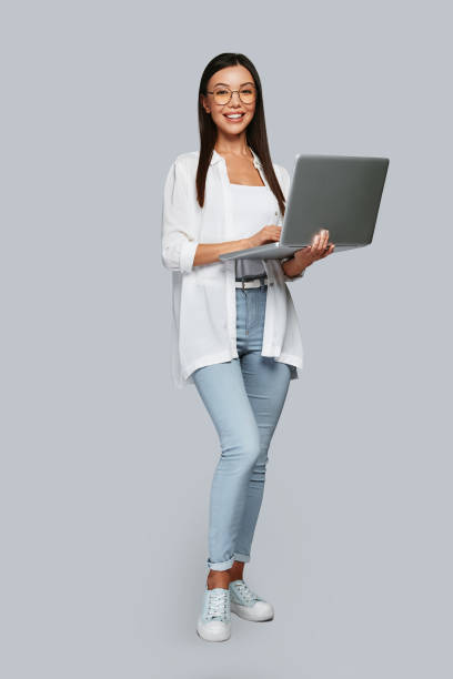 New solution every day. Full length of young Asian woman looking at camera and smiling while standing against grey background women young women standing full length stock pictures, royalty-free photos & images