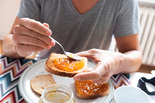 closeup of a young caucasian man, wearing a casual gray T-shirt, sitting at a set table, spreading some orange or peach jam on a toast