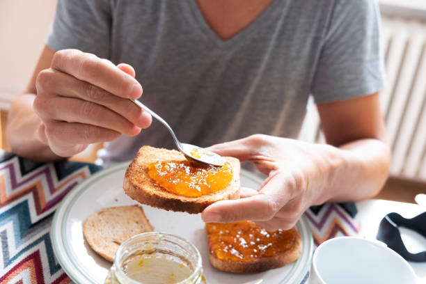 young man spreading jam on a toast closeup of a young caucasian man, wearing a casual gray T-shirt, sitting at a set table, spreading some orange or peach jam on a toast spreading stock pictures, royalty-free photos & images
