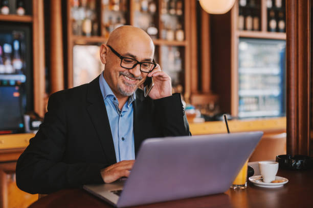 istock Bearded senior in suit using smart phone and laptop while sitting in cafeteria. On the table laptop, juice and coffee. 1073904030