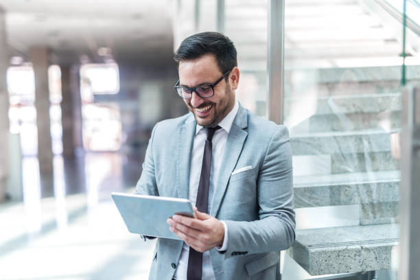 istock Smiling businessman using tablet for reading e-mail while leaning on the glass in the lobby. 1073911268