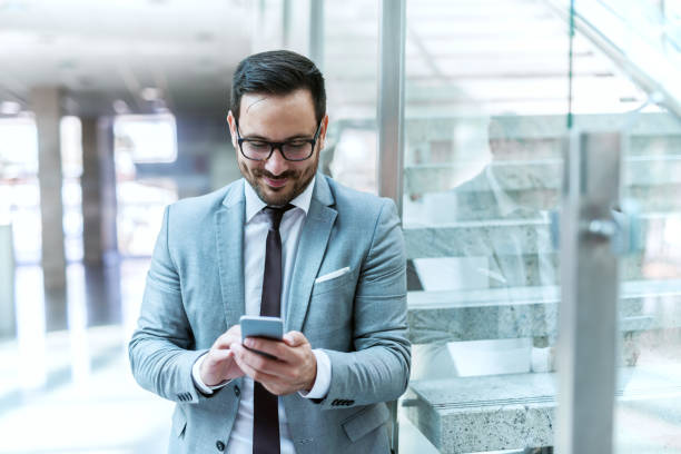 istock Happy sharp suited businessman using smart phone for writing message while leaning on the glass. 1073912454