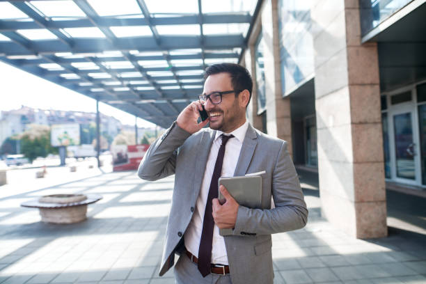 istock Smiling Caucasian businessman using smart phone and holding tablet in other hand while walking outdoors. 1073912676