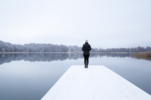 Young adult woman standing alone on edge of white snowy footbridge. Staring at lake. Unfrozen water. Early chilly morning. Peaceful atmosphere in nature. Enjoying fresh air in winter day. Back view.