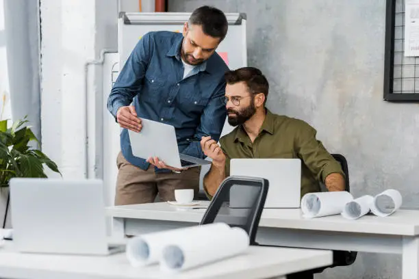 two handsome businessmen looking at laptop in office, blueprints on tabletops