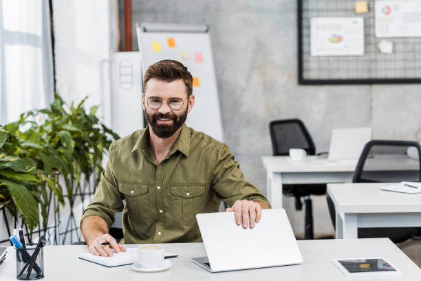 istock smiling handsome businessman sitting at table with laptop and notebook and looking at camera in office 1073952236