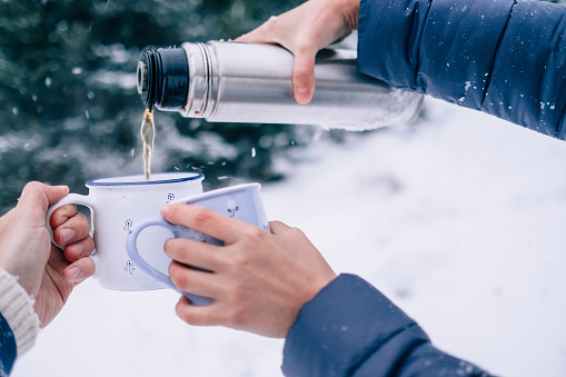 Hands with cups of tea and thermos flask in snowy winter forest close up image
