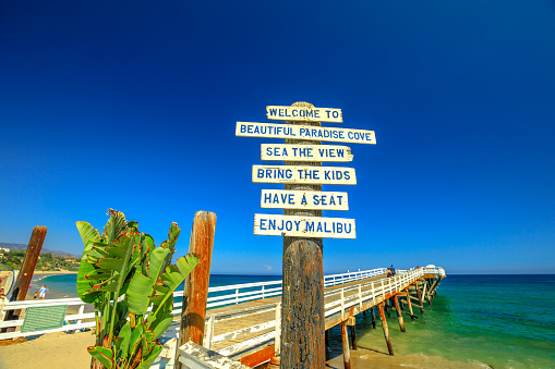 Malibu, California, United States - August 7, 2018: Paradise Cove Pier sign and wooden jetty in Paradise Cove beach, Malibu. Summer blue sky, copy space. Luxurious travel destination on Pacific Coast.
