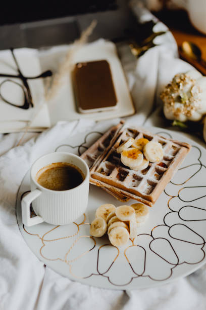placa de pequeno-almoço waffle com cobertura de banana em uma cama branca ao lado de um diário e um telefone - autumn banana breakfast close up - fotografias e filmes do acervo