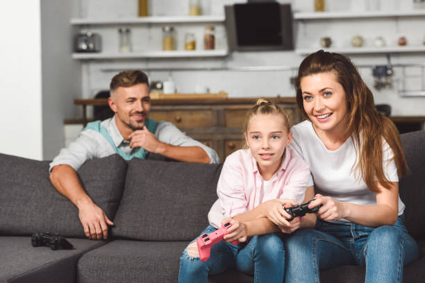 istock attractive mother and daughter playing video game while father hiding behind sofa 1074145790