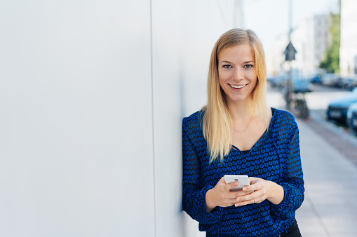 Smiling young woman holding her mobile phone as she leans against a building wall in a city street