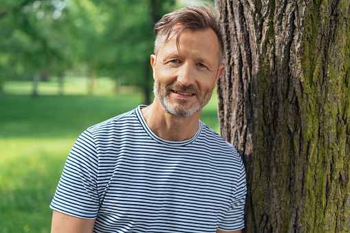 Casual middle-aged man leaning on a tree trunk in a wooded park smiling at the camera