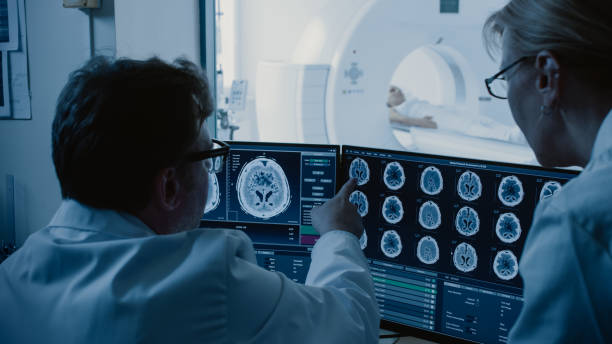 médico de sala de controle e do radiologista discutir diagnóstico enquanto procedimento assistindo e monitores mostrando resultados de exames cerebrais, em the background paciente sofre mri ou procedimento de tomografia computadorizada. - cat scan x ray medical scan x ray image - fotografias e filmes do acervo