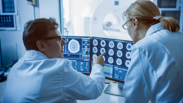 istock In Control Room Doctor and Radiologist Discuss Diagnosis while Watching Procedure and Monitors Showing Brain Scans Results, In the Background Patient Undergoes MRI or CT Scan Procedure. 1074166440