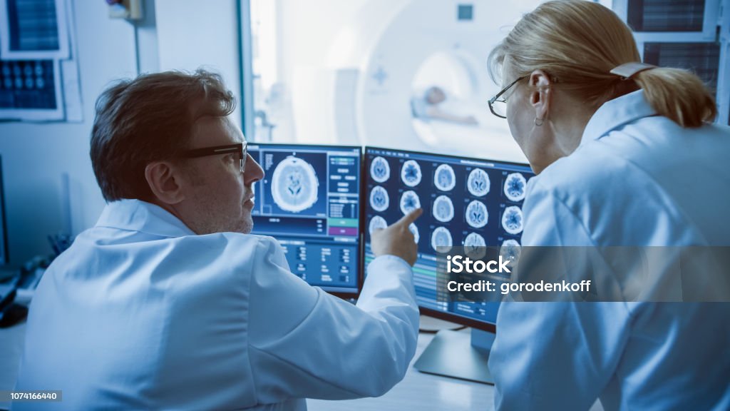 In Control Room Doctor and Radiologist Discuss Diagnosis while Watching Procedure and Monitors Showing Brain Scans Results, In the Background Patient Undergoes MRI or CT Scan Procedure. Radiotherapy Stock Photo