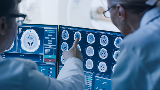 En la sala de Control médico y radiólogo discutir diagnóstico mientras ver procedimiento y monitores que muestran resultados de las exploraciones del cerebro, en el fondo paciente se somete a resonancia magnética o tomografía computarizada. photo