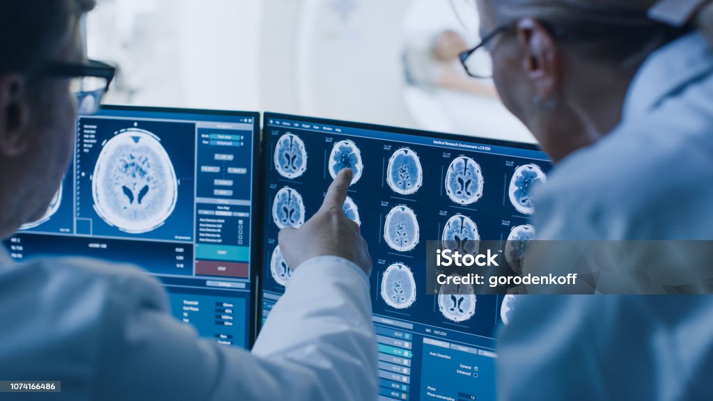 In Control Room Doctor and Radiologist Discuss Diagnosis while Watching Procedure and Monitors Showing Brain Scans Results, In the Background Patient Undergoes MRI or CT Scan Procedure. Cancer - Illness Stock Photo