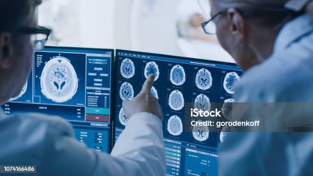 In Control Room Doctor And Radiologist Discuss Diagnosis While Watching Procedure And Monitors Showing Brain Scans Results In The Background Patient Undergoes Mri Or Ct Scan Procedure Stock Photo - Download Image Now