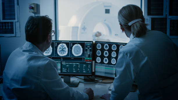 médico de sala de controle e do radiologista discutir diagnóstico enquanto procedimento assistindo e monitores mostrando resultados de exames cerebrais, em the background paciente sofre mri ou procedimento de tomografia computadorizada. - radiologist - fotografias e filmes do acervo