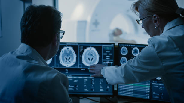 istock In Control Room Doctor and Radiologist Discuss Diagnosis while Watching Procedure and Monitors Showing Brain Scans Results, In the Background Patient Undergoes MRI or CT Scan Procedure. 1074166616