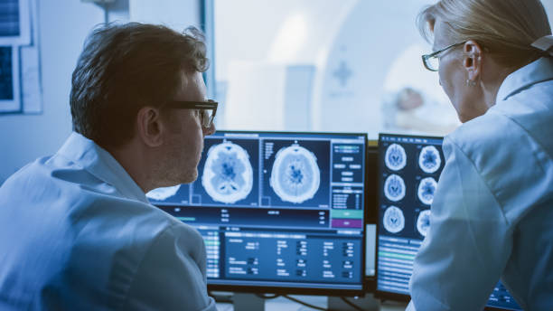 istock In Control Room Doctor and Radiologist Discuss Diagnosis while Watching Procedure and Monitors Showing Brain Scans Results, In the Background Patient Undergoes MRI or CT Scan Procedure. 1074166634