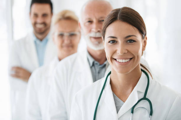 Group of doctors looking at the camera. Closeup front view of group of mixed age doctors at a hospital standing in a row and smiling at the camera. Mid 20's intern is in foreground with the rest of the team out of focus. assistant photos stock pictures, royalty-free photos & images