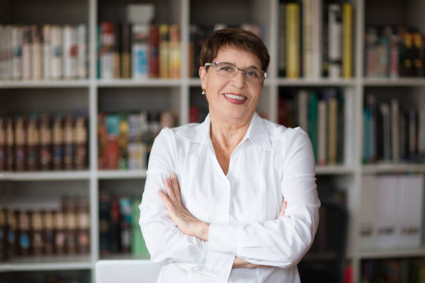 Portrait of  senior businesswoman wearing glasses head shot in a white shirt, crossed hands looking at the camera with a warm friendly smile against the background of a bookcase stock photo