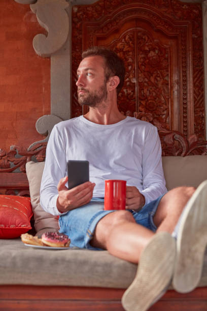 istock Man sitting on a terrace sofa and drinking coffee/tea, eating donuts while using cellphone. 1074210732