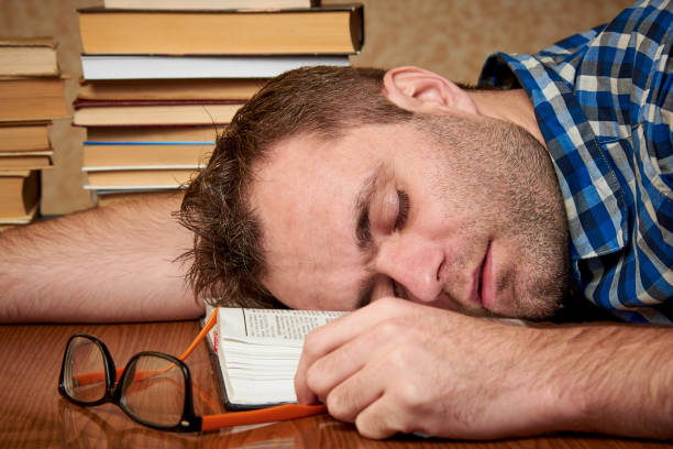 istock A tired and tortured disheveled student with glasses is sleeping at a table with stacks of books. 1074219068
