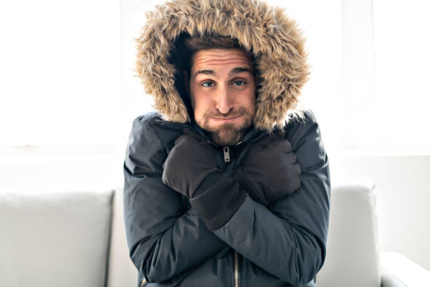 un hombre frío en el sofá en casa con abrigo de invierno - frío fotografías e imágenes de stock