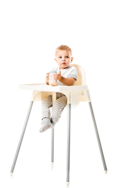 adorable toddler boy holding baby bottle and sitting in highchair isolated on white adorable toddler boy holding baby bottle and sitting in highchair isolated on white high chair stock pictures, royalty-free photos & images