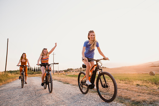 Three young women are racing on bikes along a dirt road in Tuscany, Italy.