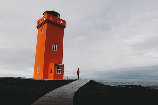Silhouette of man in red shirt staying near remote beautiful orange lighthouse on Snaefellsnes peninsula in Iceland