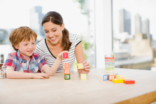 Positive kids psychology. Professional female therapist with notepad watching little girl playing with colorful blocks, sitting at table in office and smiling at each other