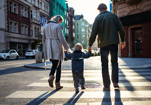 A rear view of small toddler boy with parents crossing a road outdoors in city, holding hands.