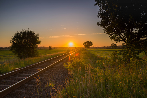 Railroad track through fields with trees at sunset. Lens flares. Near Jever, Friesland - district, Lower Saxony, Germany, Europe.